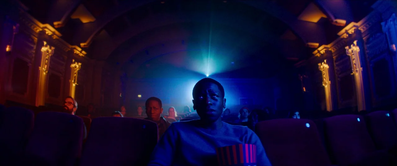 Rye Lane (2023) movie still: medium — A dimly lit movie auditorium with rows of red velvet seats filled with people. The central character sits in…