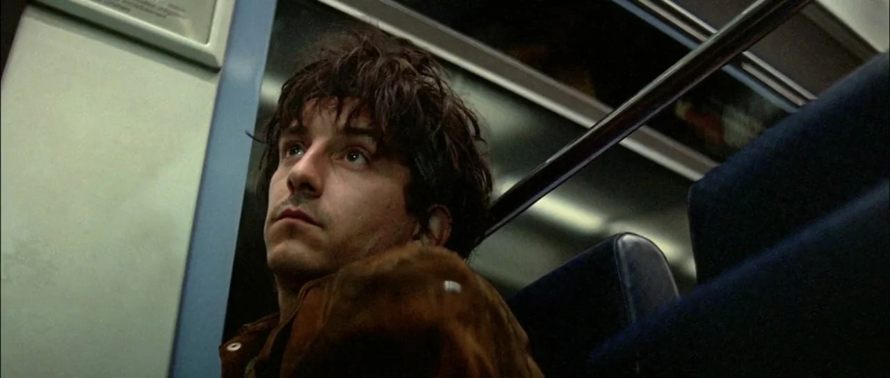 Subway (1985) movie still: closeup — The interior of a late-night subway car features dark blue padded seats and windows. A young man with messy…