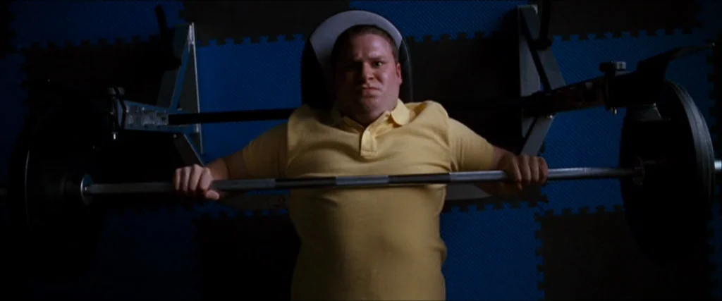 Observe And Report (2009) movie still: medium — A man lies on a bench press station with blue and black foam flooring behind. He is lifting a metal…