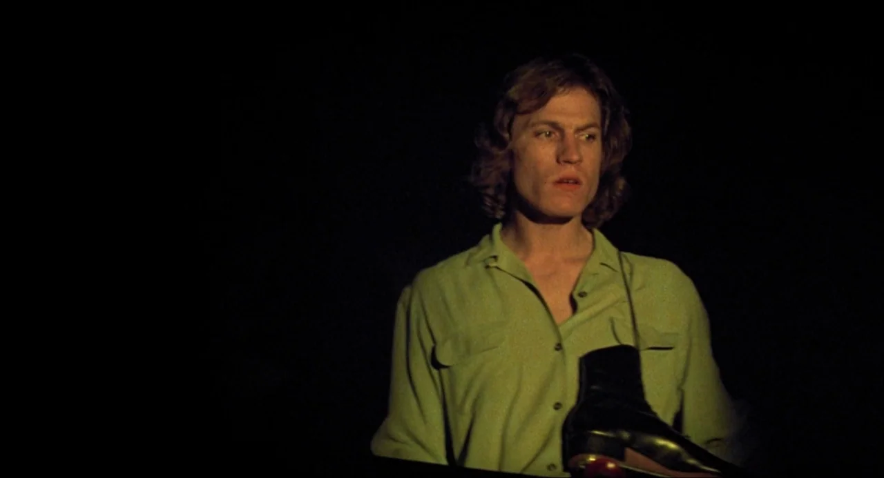 Xanadu (1980) movie still: medium — A man stands centered against a completely dark, featureless backdrop. He wears a green button-down shirt and…