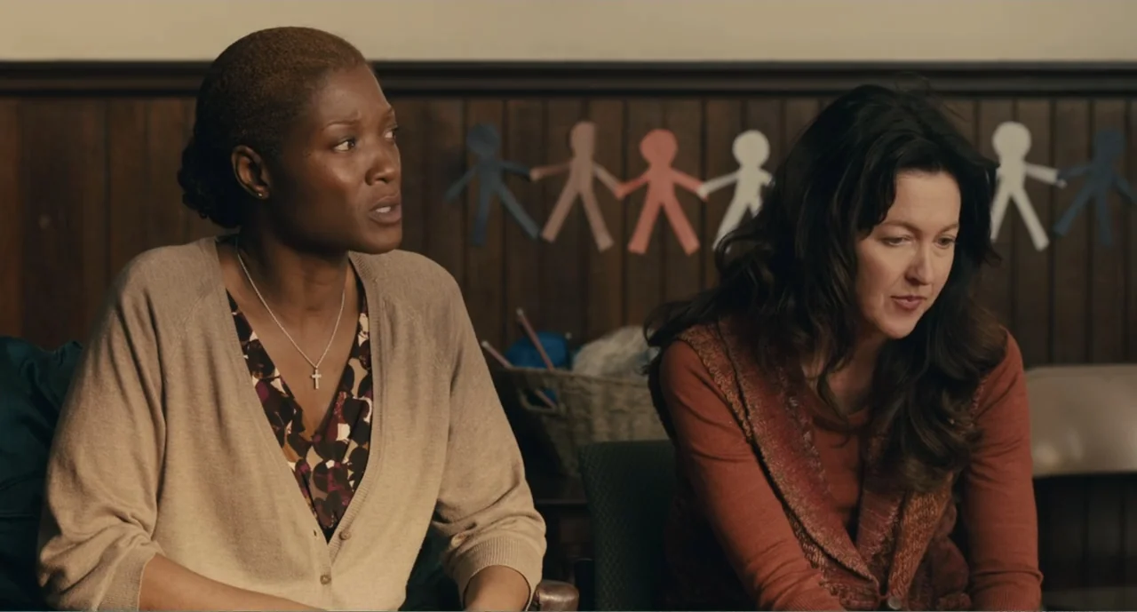 Lila & Eve (2015) movie still: medium — A community center interior features wood paneling and paper doll cutouts on the wall. Two women sit…
