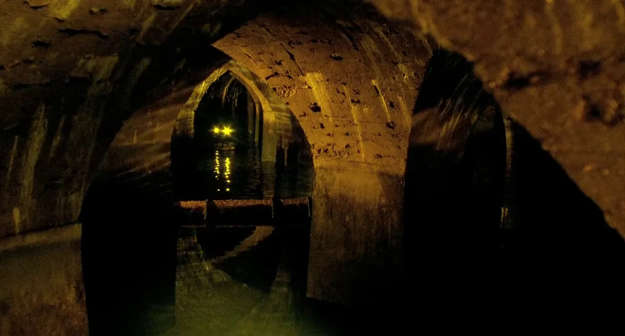 Delicatessen (1991) movie still: establishing — An underground stone tunnel features arched passageways and a pool of dark water. Two bright yellow…
