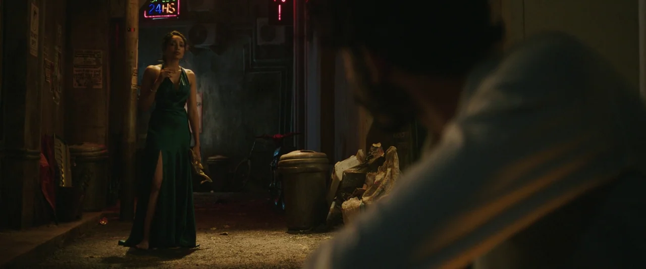 Monkey Man (2024) movie still: fullbody — A dark, narrow urban alleyway features litter bins and discarded clutter under red neon signage. A woman…