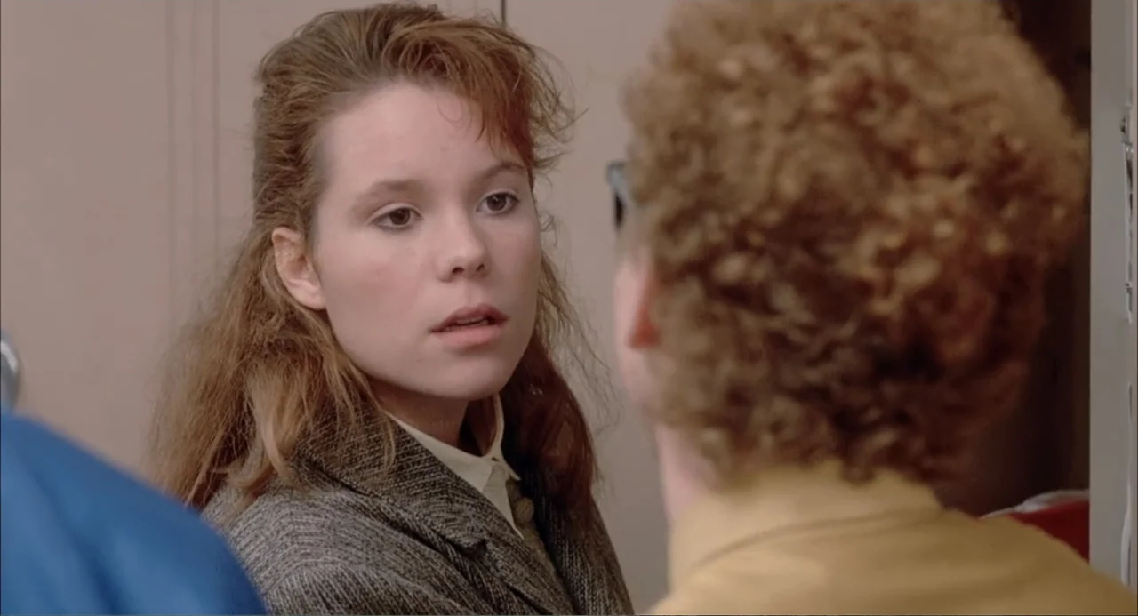 Teen Witch (1989) movie still: medium — A high school hallway with beige lockers visible in the background. A young woman in a tweed blazer looks…
