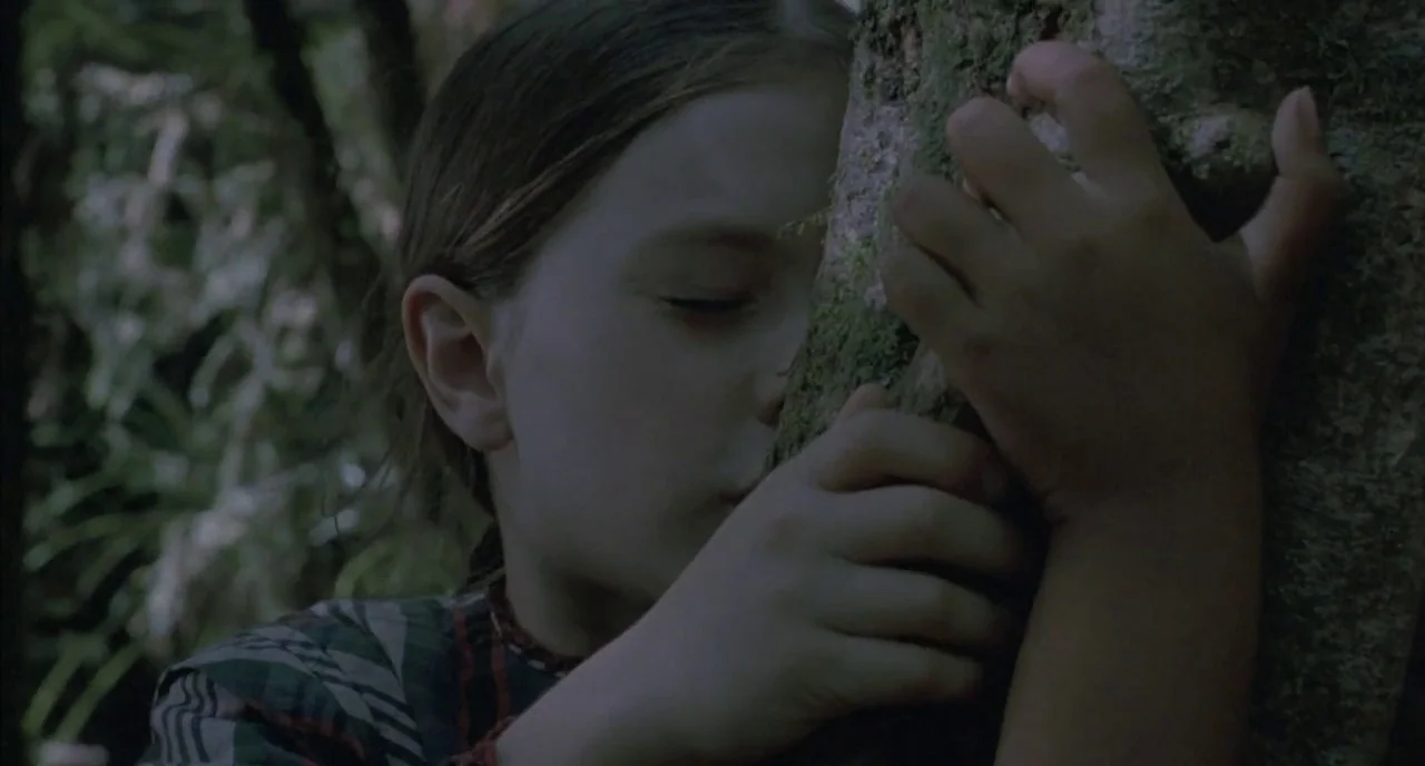 The Piano (1993) movie still: closeup — A dense patch of woodland with textured, moss-covered tree trunks and shadowed undergrowth. A young girl…