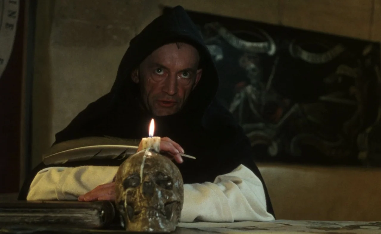 The Pit and The Pendulum (1991) movie still: medium — A dimly lit stone chamber contains a wooden table cluttered with medieval manuscripts. A monk…
