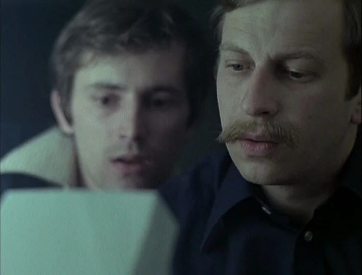 Camera Buff (1979) movie still: medium — An dimly lit interior workspace with muted walls and minimal background detail. Two men lean forward…