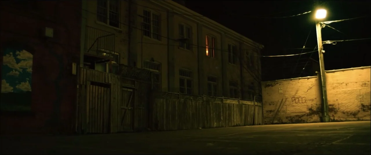 I'll Take Your Dead (2018) movie still: wide — An exterior alleyway at night features a brick building beside a tall wooden fence. A single bright…