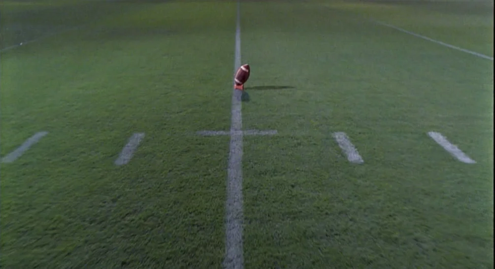 Raising Arizona (1987) movie still: establishing — An expanse of green grass with painted white yard lines on a football field. A single football…