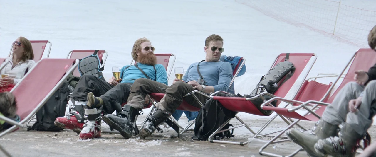 Force Majeure (2014) movie still: wide — A snowy outdoor terrace at a ski resort features several rows of red lounge chairs. Three people sit in the…
