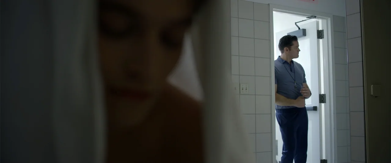 Init!ation (2021) movie still: medium — A tiled locker room with a bright doorway leading to an exterior space. A man in athletic wear stands in the…