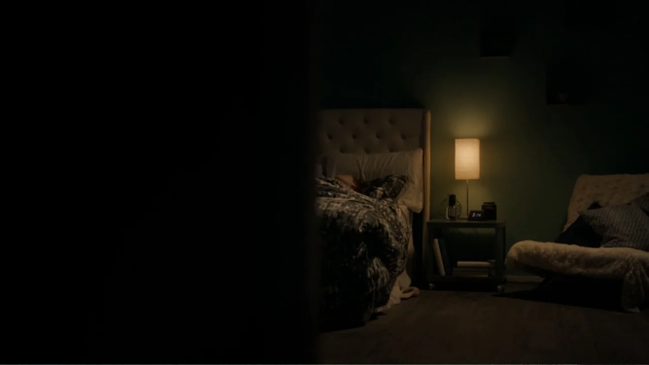 Sightless (2017) movie still: medium — A dark bedroom features a bed with a patterned duvet and a bedside lamp. The glowing lamp illuminates a small…