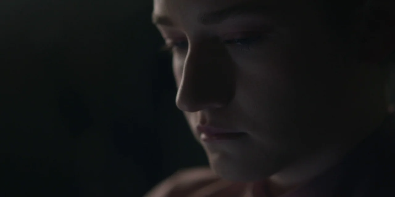 The Assistant (2019) movie still: closeup — A dark, underexposed interior space surrounds the subject. A young woman looks down with her face…