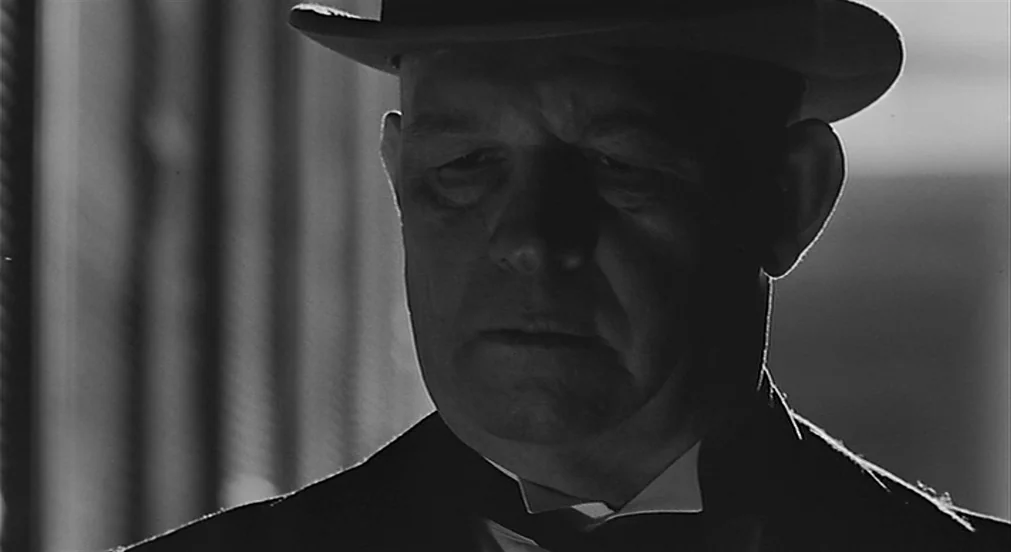 Kafka (1991) movie still: closeup — A man wearing a hat is lit with high contrast side lighting. He gazes downward with a neutral facial expression.