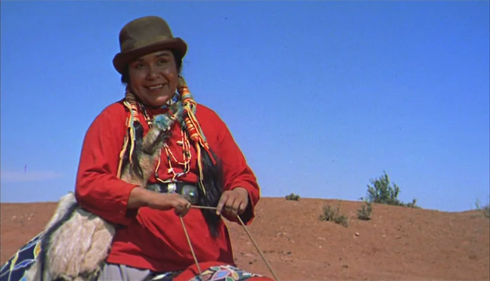 The Searchers movie still: medium — An arid, sun-drenched desert landscape with sparse scrub brush under a clear blue sky. A woman in a red shirt…