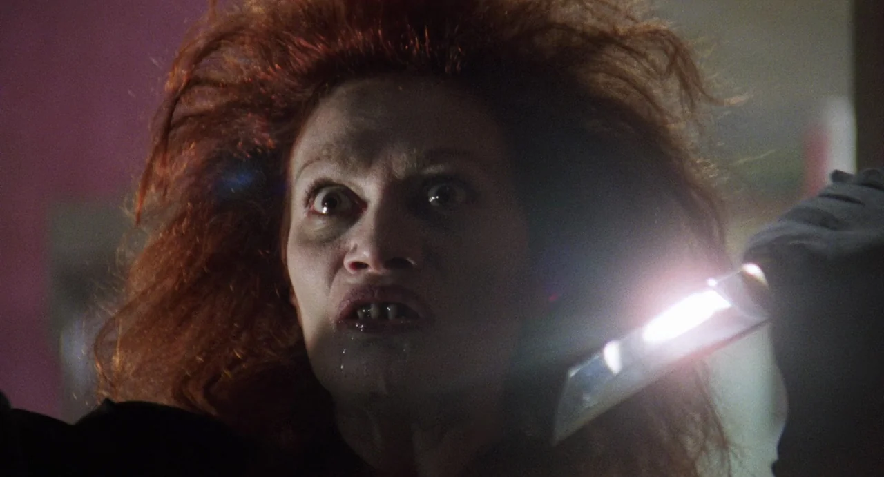 The Spider Labyrinth (1988) movie still: closeup — A dark, dimly lit environment with indistinct background shadows. A woman with wild red hair…