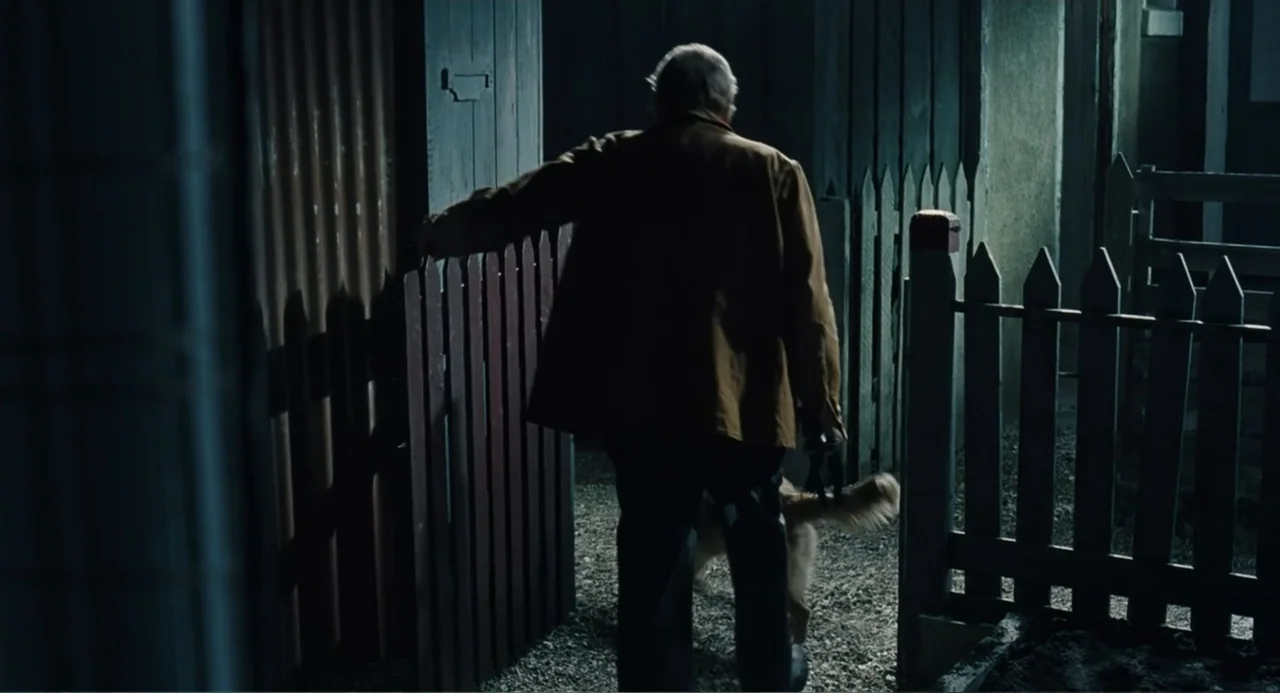 Le Havre (2011) movie still: fullbody — A wooden fenced enclosure stands in a dark exterior environment at night. An elderly man walks through the…