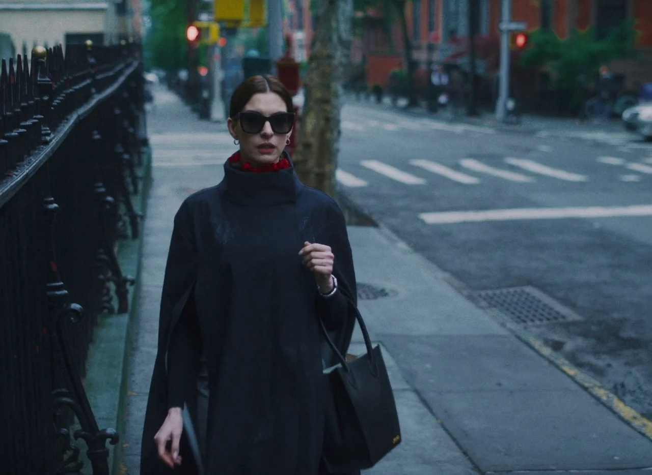 She Came To Me (2023) movie still: medium — A woman walks along a paved sidewalk lined with an ornate black metal fence. The background shows a…