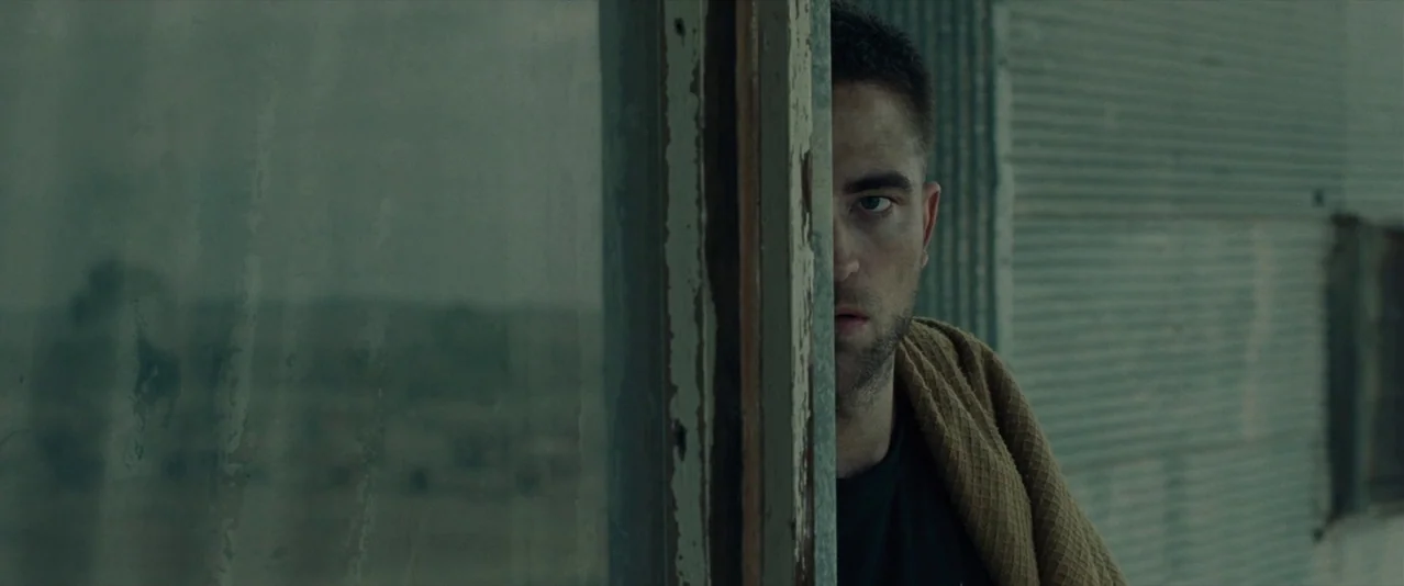 The Rover (2014) movie still: closeup — A man looks from behind a wooden frame attached to a corrugated metal wall. The exterior environment is…