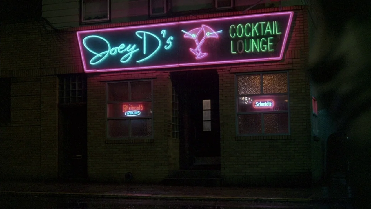 Baby It’s You (1983) movie still: establishing — A brick building facade features a prominent neon sign for a cocktail lounge. Two smaller neon…