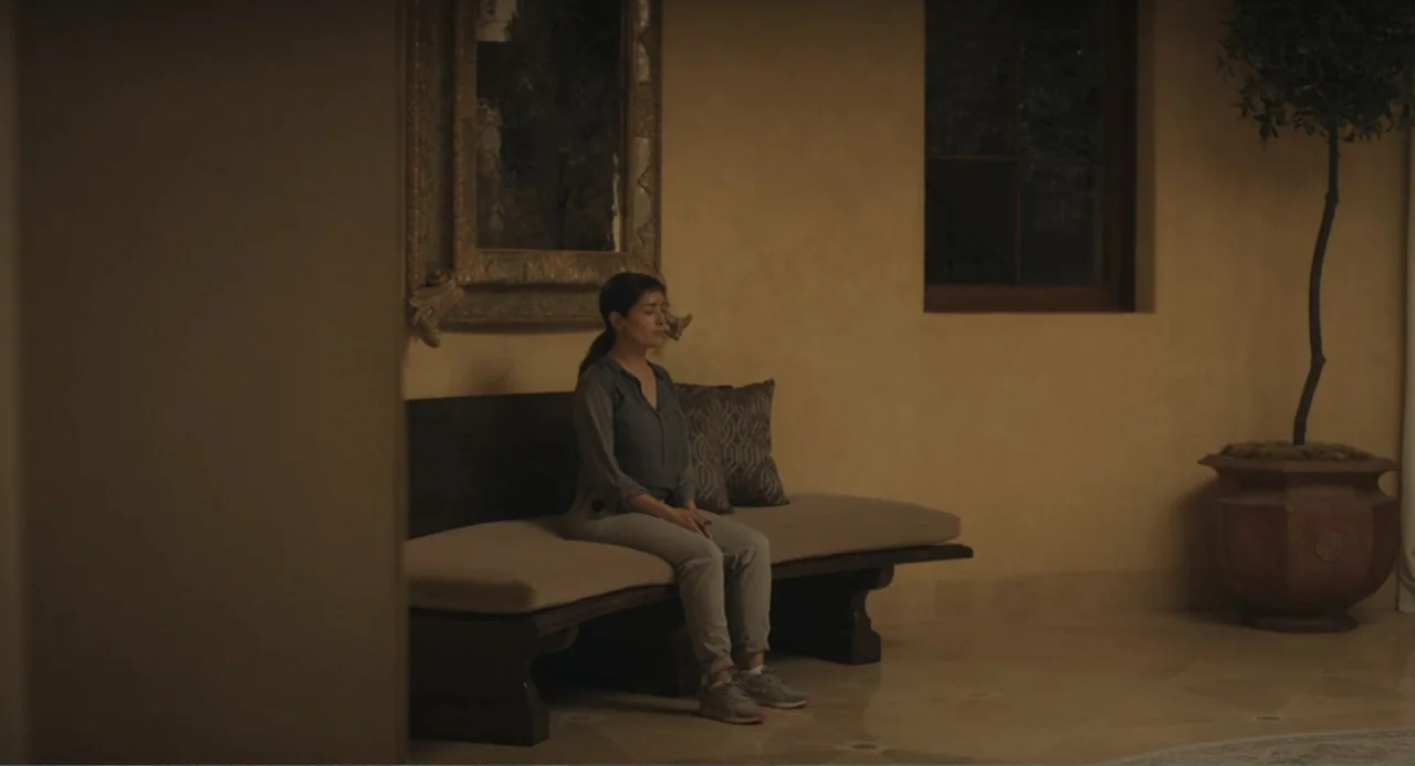 Beatriz at Dinner (2017) movie still: fullbody — An interior space features a long bench against a tan wall with a mirror. A woman sits on the bench…