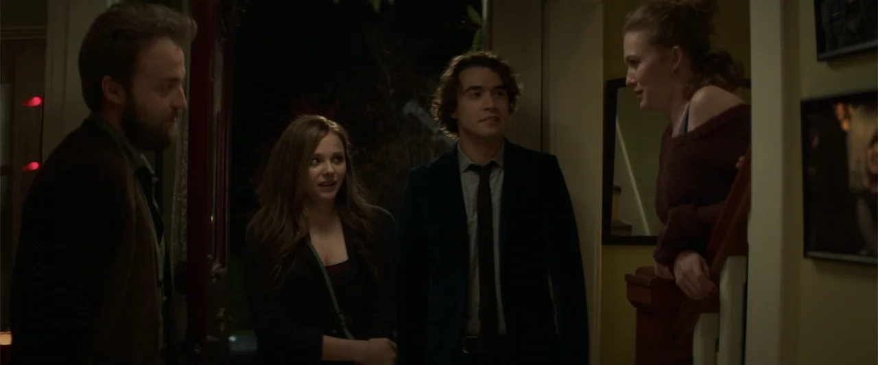 If I Stay (2014) movie still: medium — A dimly lit residential hallway at night with framed pictures on the walls. Three young adults face a woman…
