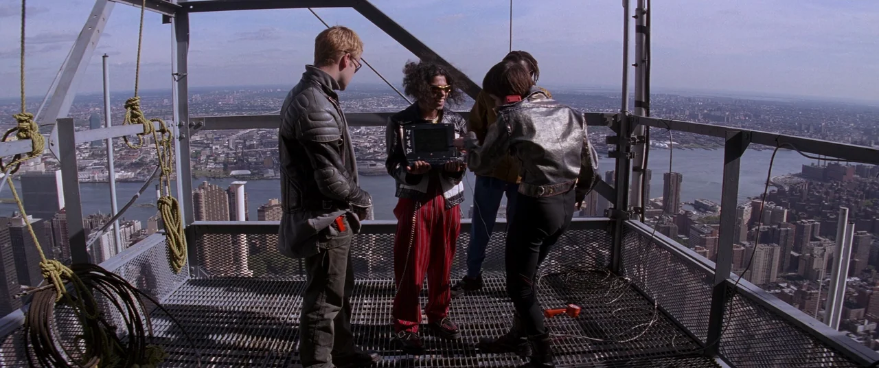 Hackers (1995) movie still: fullbody — A high-altitude open-grated metal maintenance platform overlooking a vast sprawling city harbor area. Four…