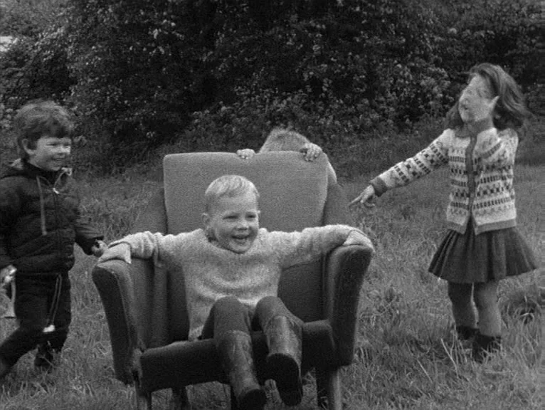 Cathy Come Home (1969) movie still: fullbody — A group of children play with a discarded armchair in a grassy field. The boy sits in the chair while…