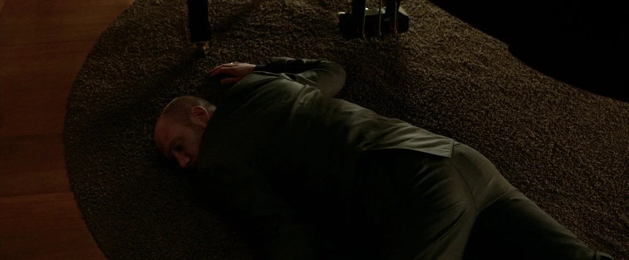 Borgman (2013) movie still: medium — A man in a suit lies motionless on a circular brown textured rug. His body is positioned near the legs of a…