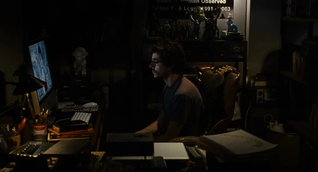 Sidewalls (2011) movie still: medium — A dark, cluttered room featuring a man sitting at a desk facing a computer. The background contains shelves…