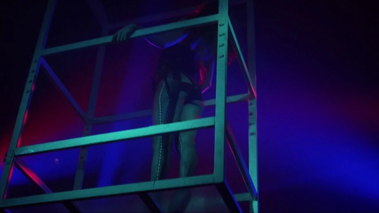 Bad Lieutenant (1992) movie still: medium — A dark, neon-lit environment contains a large, skeletal metal cage structure. A woman wearing a leotard…