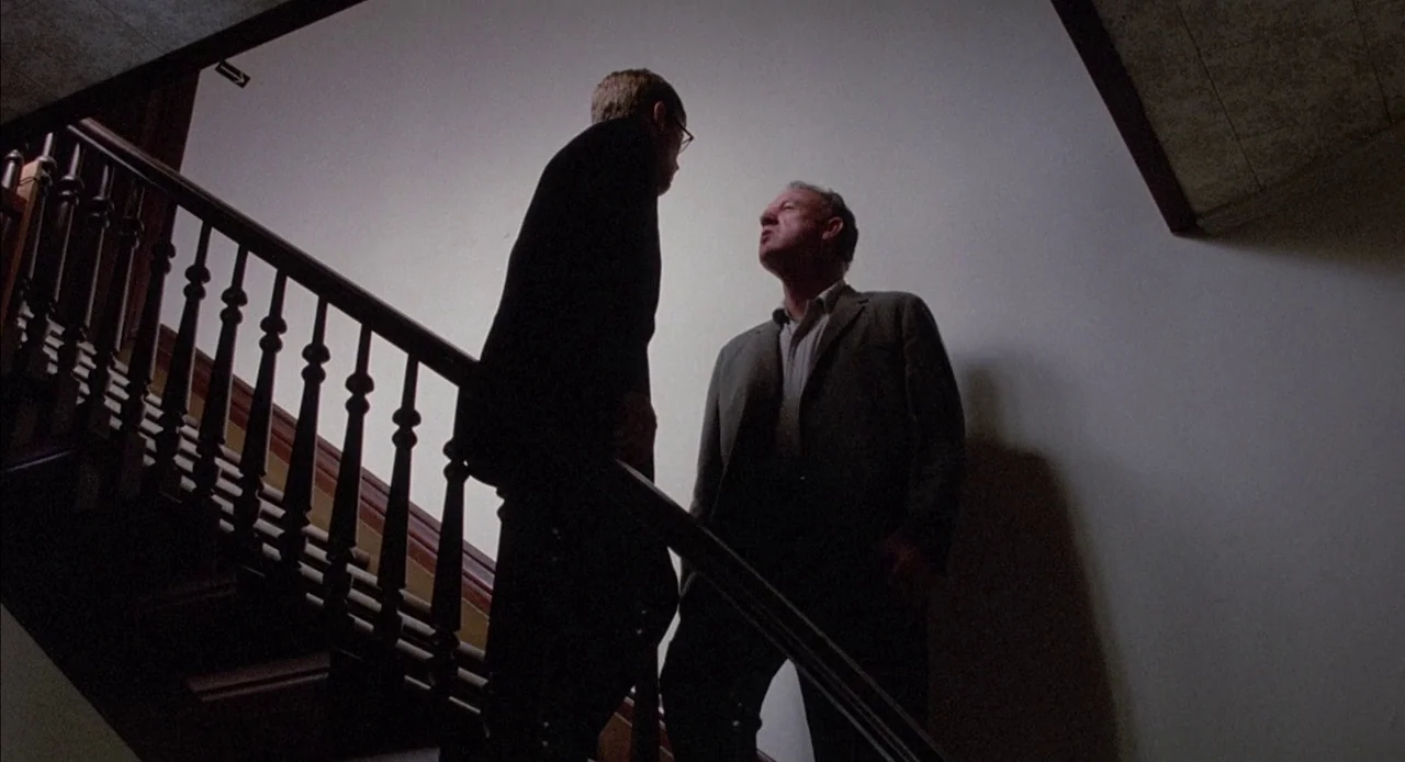 Mississippi Burning (1988) movie still: fullbody — Two men stand on a wooden staircase with decorative railings against a plain wall. The taller man…