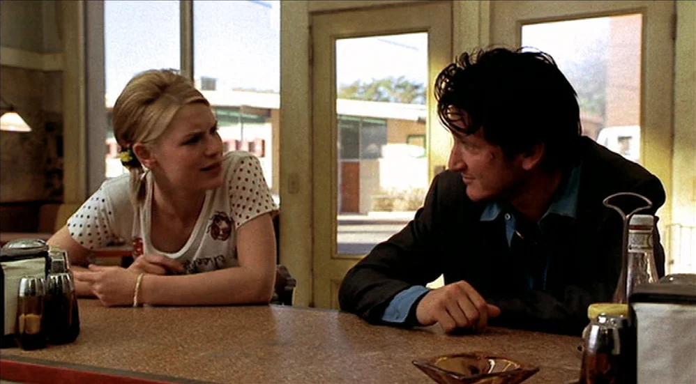 U-Turn (1997) movie still: medium — A sunlit diner interior with large glass windows and worn surfaces. A young woman and a man in a suit sit at the…