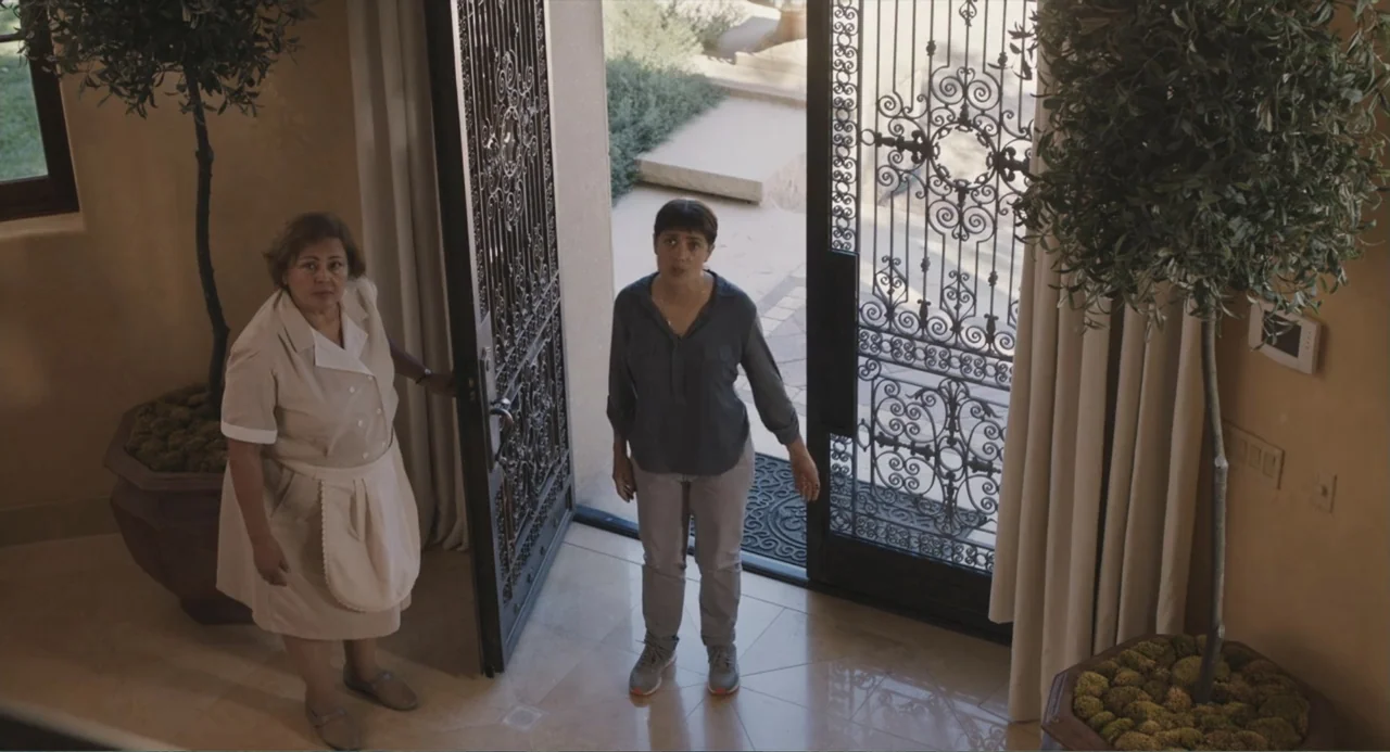 Beatriz at Dinner (2017) movie still: fullbody — A spacious home entryway features a large ornamental iron door and potted indoor trees. Two women…