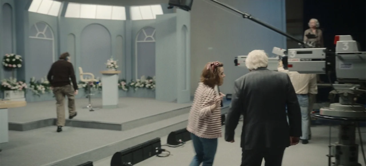 One Life (2023) movie still: medium — A television studio features a raised set with arched panels and floral arrangements. A man and a woman walk…