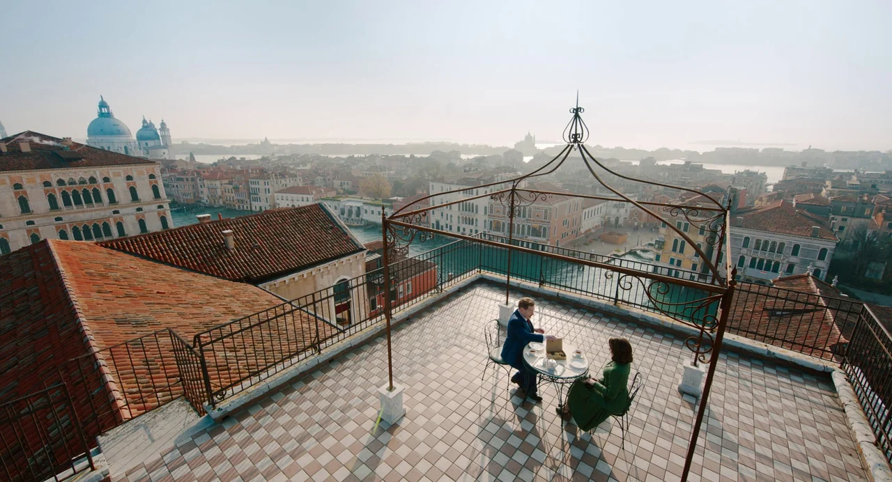 A Haunting In Venice (2023) movie still: wide — A tiled rooftop terrace overlooks a canal city with historic buildings and distant domes. A man and…
