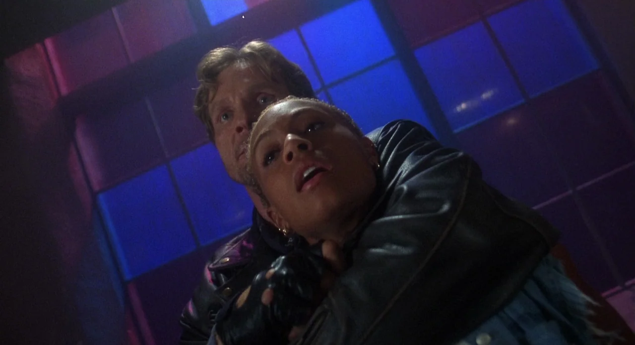 Tales from the Crypt: Demon Knight (1995) movie still: medium — A dark industrial interior features a paneled grid window backlit by intense blue…