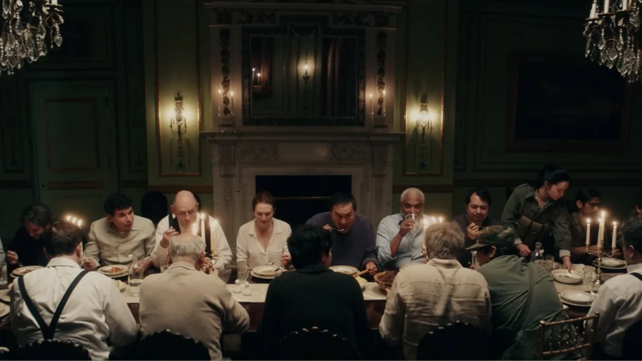 Bel Canto (2018) movie still: wide — A formal dining room features light green paneled walls and a large fireplace mantelpiece. A group of adults is…
