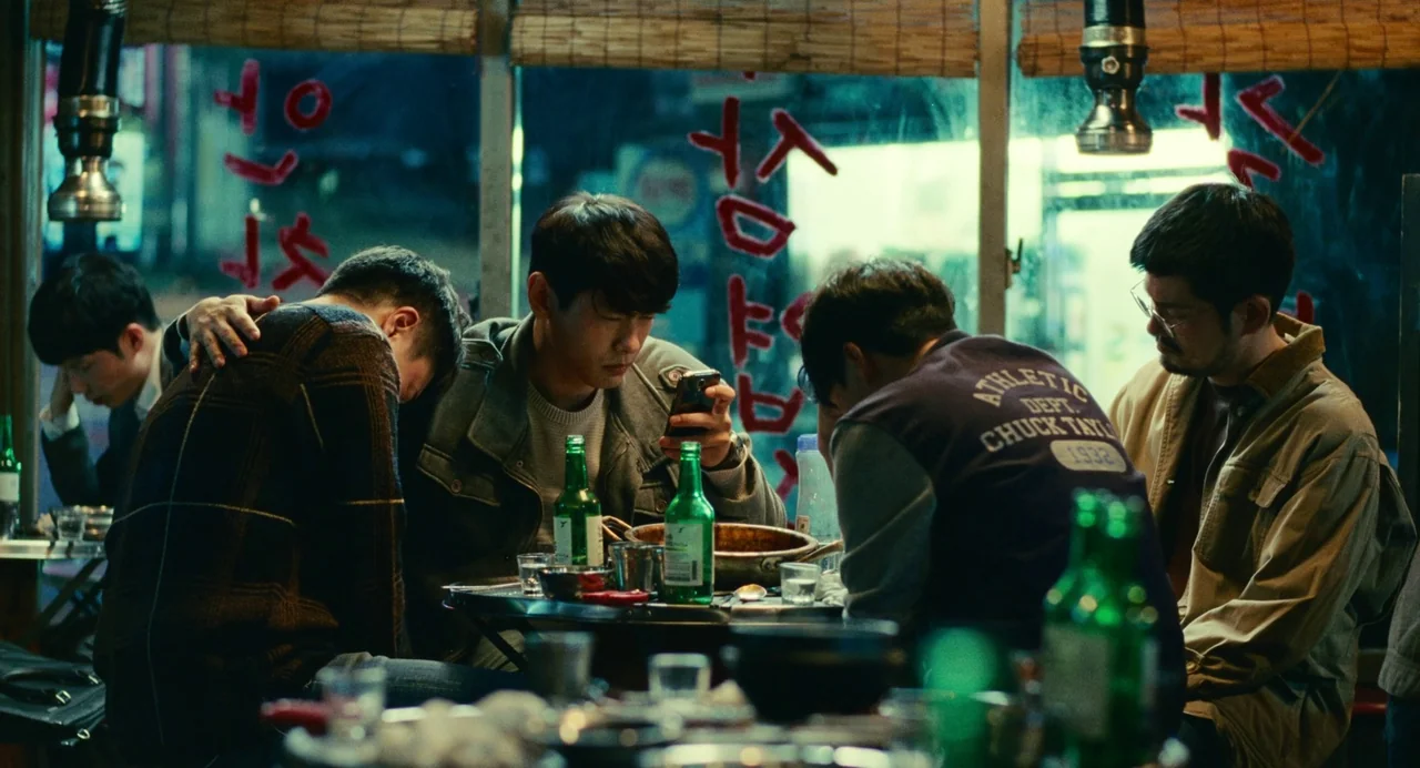 Past Lives (2023) movie still: medium — A small outdoor-style restaurant with red Korean text written on the window glass. Four young men sit at a…