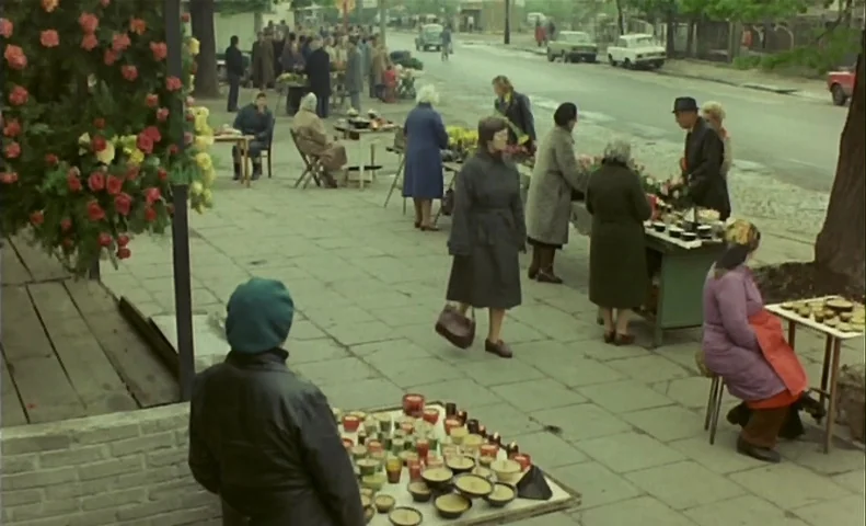 No End (1985) movie still: wide — A sidewalk outdoor market features multiple stalls with candles and small goods for sale. Pedestrians walk along…