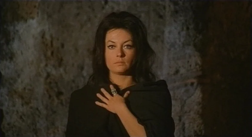 Kill, Baby Kill (1966) movie still: medium — A woman stands in front of a textured, rough stone wall in dim light. She holds a dark shawl over her…