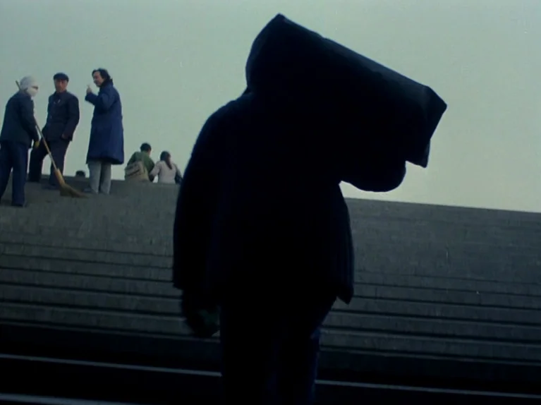 Black Snow (1990) movie still: fullbody — Wide, concrete exterior stairs ascend toward an overcast sky with muted natural light. A dark silhouette…