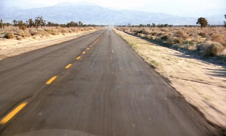 The Brown Bunny (2003) movie still: establishing — A flat two-lane asphalt road runs straight through a vast arid desert plain. Scrubby vegetation…