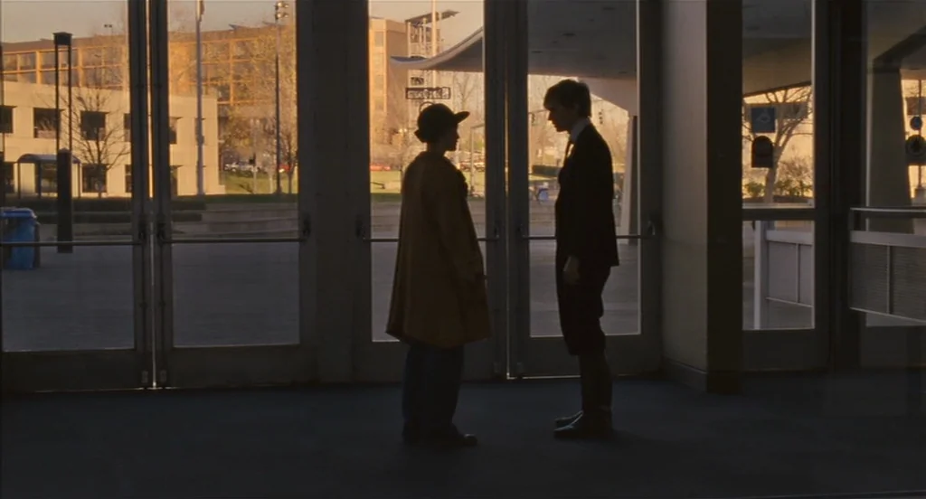 Restless (2011) movie still: fullbody — A glass-fronted building entryway faces a sunlit paved plaza during late afternoon. Two people stand in…