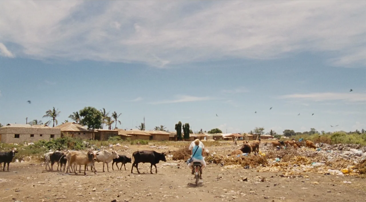 Paradise: Love (2012) movie still: wide — A dry, dusty landscape is scattered with trash and grazing cattle near small buildings. A woman in a light…