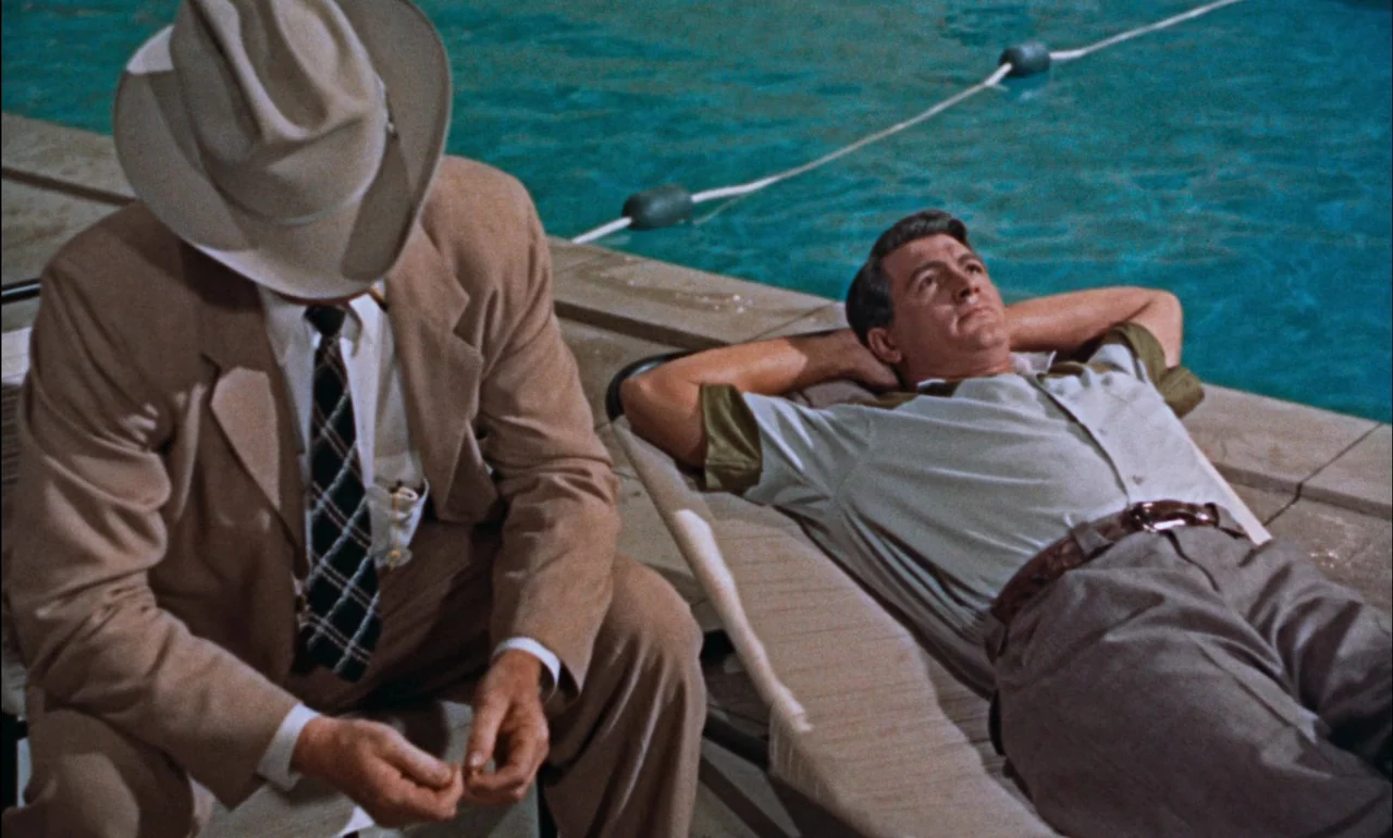 Giant (1956) movie still: medium — A concrete pool deck edge sits adjacent to deep blue water with floating buoys. One man in a suit sits beside…