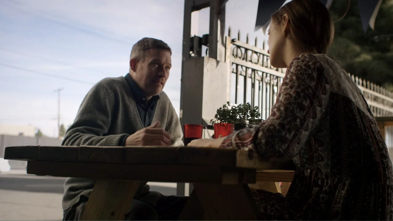 Totem (2022) movie still: medium — An outdoor seating area features a weathered wooden table with red plastic cups. A man and a woman sit facing…