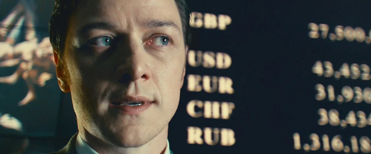 Trance (2013) movie still: closeup — A dark room features a large digital screen displaying currency exchange rates in gold text. A man stands…