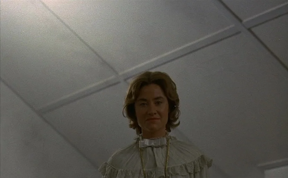 To The Devil a Daughter (1976) movie still: medium — A woman stands in a pale white hallway with panelled ceiling tiles above her. She looks down…