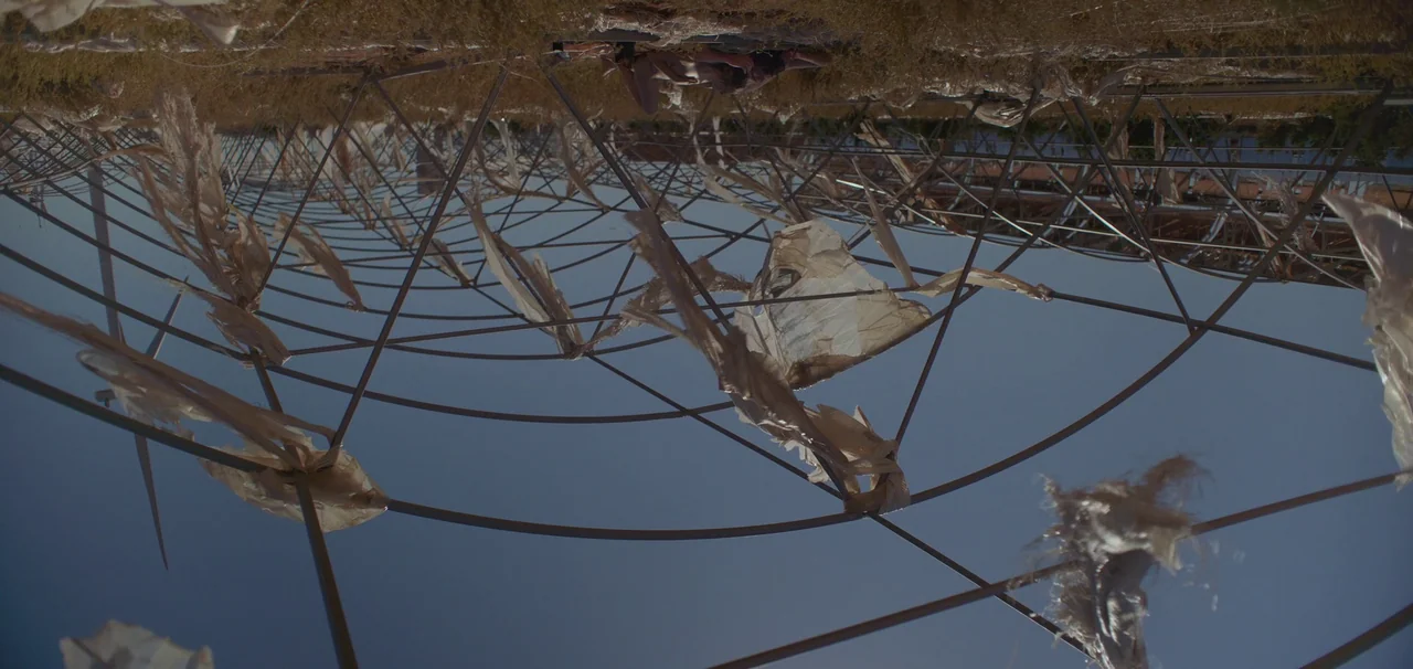 Foe (2023) movie still: wide — A metal geodesic framework sits in a field covered with tattered, flapping plastic sheets. The camera angle is…